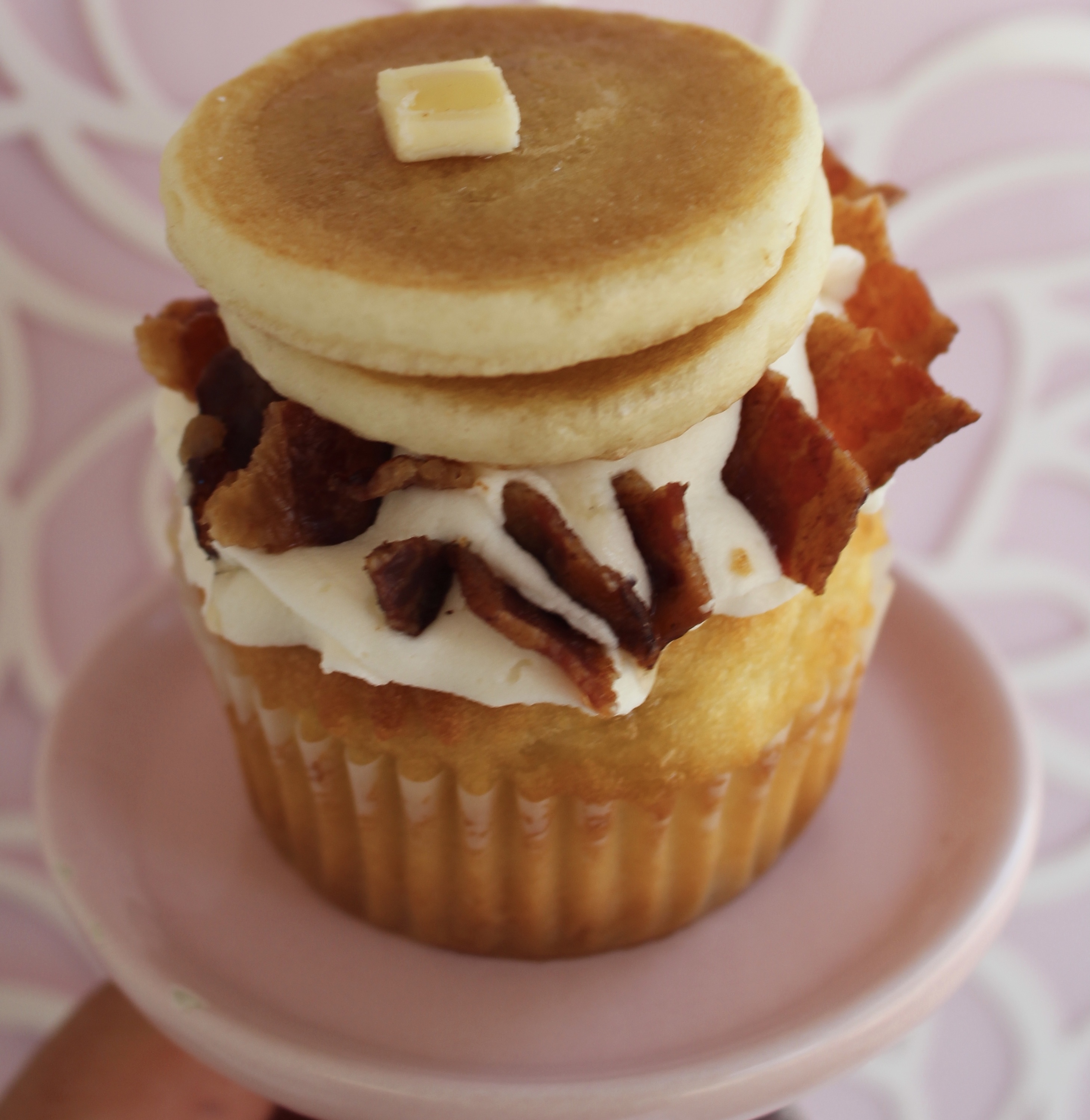 Maple cake with a vanilla buttercream topping, bacon pieces, a mini stack of pancakes, and drizzled with maple syrup!